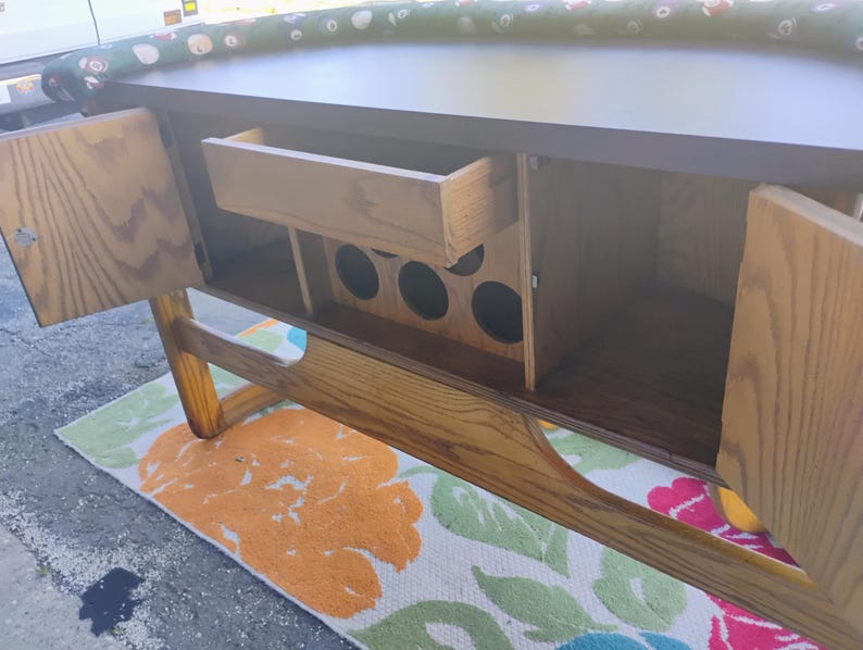 May include: A wooden cabinet with open doors, revealing a drawer and storage space. The cabinet has a dark brown top and light brown wooden doors and frame. The interior includes circular cutouts, likely for bottles. A colorful floral rug is visible below.