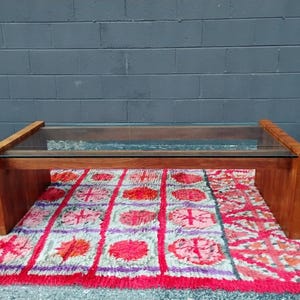 May include: A rectangular coffee table with a clear glass top and a wooden frame. The table is supported by a solid wooden base. The table is placed on a colorful rug with red, pink, and purple patterns.