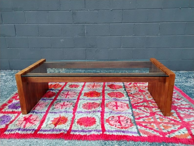 May include: A rectangular coffee table with a clear glass top and a wooden frame. The table is supported by a solid wooden base. The table is placed on a colorful rug with red, pink, and purple patterns.