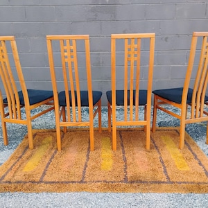 May include: Four wooden chairs with high, slatted backrests featuring a grid pattern at the top. Each chair has a black upholstered seat and four straight legs. The chairs are arranged in a row on a rectangular, shaggy, orange and brown rug.
