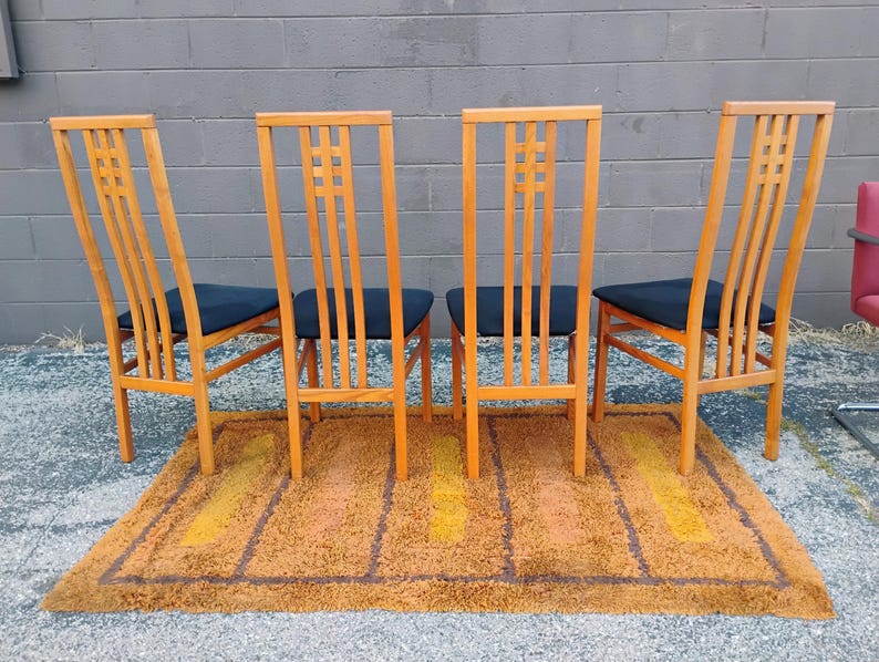 May include: Four wooden chairs with high, slatted backrests featuring a grid pattern at the top. Each chair has a black upholstered seat and four straight legs. The chairs are arranged in a row on a rectangular, shaggy, orange and brown rug.