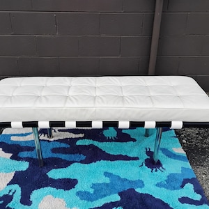 May include: A white leather bench with a tufted design, black and white striped support, and chrome legs. The bench is placed on a blue and white patterned rug. The bench measures approximately 60 inches long.