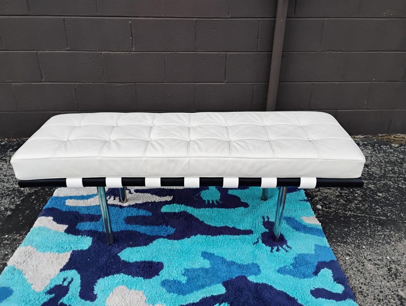May include: A white leather bench with a tufted design, black and white striped support, and chrome legs. The bench is placed on a blue and white patterned rug. The bench measures approximately 60 inches long.