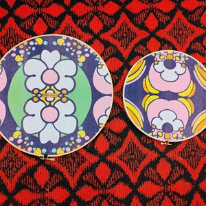 May include: Two round embroidery hoops with floral designs. The larger hoop features a blue background with white, pink, and yellow flowers. The smaller hoop has a similar design. Both hoops have wooden frames and are set against a red patterned background.