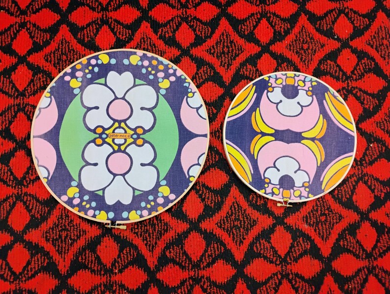 May include: Two round embroidery hoops with floral designs. The larger hoop features a blue background with white, pink, and yellow flowers. The smaller hoop has a similar design. Both hoops have wooden frames and are set against a red patterned background.
