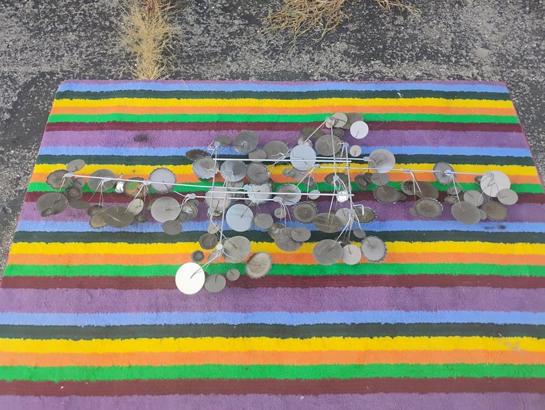 May include: A wall sculpture featuring a horizontal metal frame with numerous thin wire stems extending outward. Attached to these stems are various circular metal discs in shades of silver, gray, and muted brown, creating a layered, branching effect.