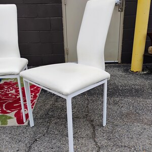 May include: A white upholstered chair with a curved back and a square seat. The chair has a white metal frame. The chair is outdoors, with a dark brick wall and a yellow pole in the background.