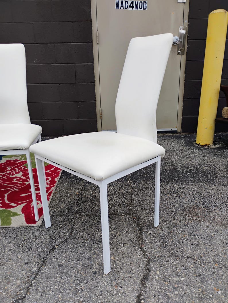 May include: A white upholstered chair with a curved back and a square seat. The chair has a white metal frame. The chair is outdoors, with a dark brick wall and a yellow pole in the background.