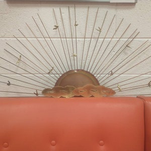May include: A gold metal wall sculpture depicting a stylised sunrise with birds flying in the sky. The sculpture has a copper-coloured sun with clouds and a fan of thin metal rays.