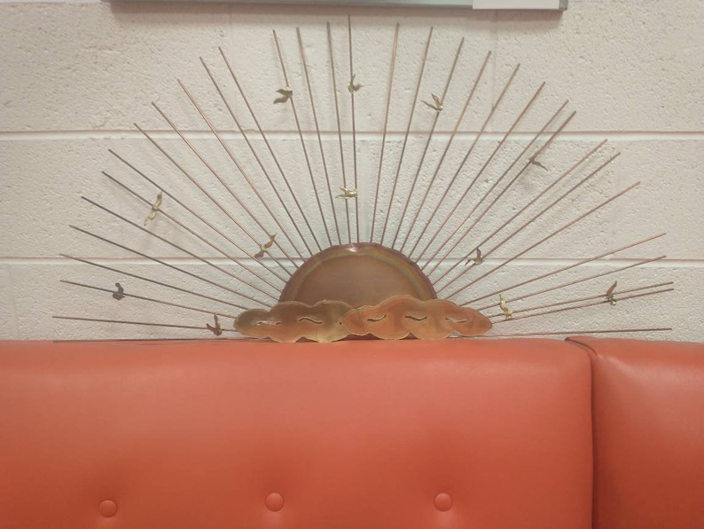 May include: A gold metal wall sculpture depicting a stylised sunrise with birds flying in the sky. The sculpture has a copper-coloured sun with clouds and a fan of thin metal rays.