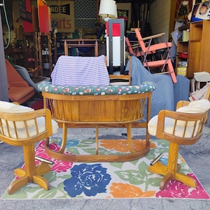 May include: A wooden baby cradle with a floral patterned rim and a striped pillow, flanked by two wooden swivel chairs with cream-colored cushions. The chairs and cradle sit on a floral patterned rug.