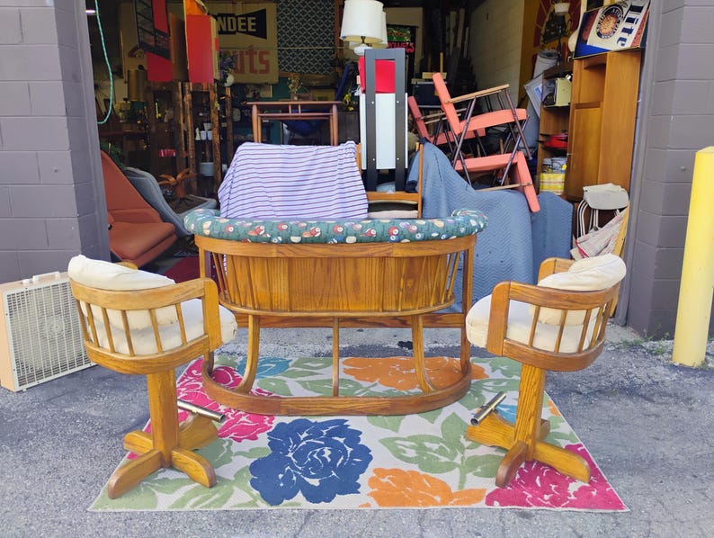 May include: A wooden baby cradle with a floral patterned rim and a striped pillow, flanked by two wooden swivel chairs with cream-colored cushions. The chairs and cradle sit on a floral patterned rug.