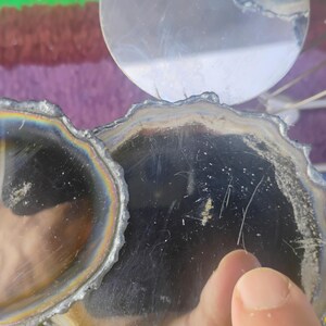 May include: Two circular coasters with dark, reflective surfaces and irregular, textured edges. The coasters feature a metallic, iridescent sheen around the perimeter, creating a rainbow-like effect against the deep black centers of the circular forms.