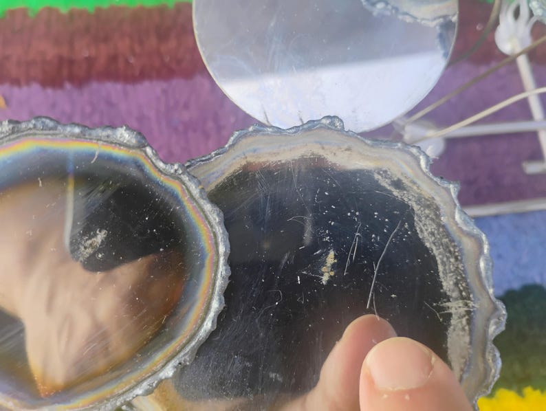 May include: Two circular coasters with dark, reflective surfaces and irregular, textured edges. The coasters feature a metallic, iridescent sheen around the perimeter, creating a rainbow-like effect against the deep black centers of the circular forms.