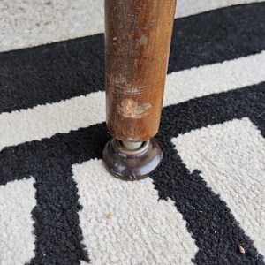 Puede incluir: Una pata de mueble cil&iacute;ndrica de madera con un rasgu&ntilde;o visible y un acabado desgastado cerca de la base, unida a un deslizador de suelo o tapa de pl&aacute;stico de color marr&oacute;n oscuro. La pata est&aacute; colocada verticalmente, mostrando la veta.