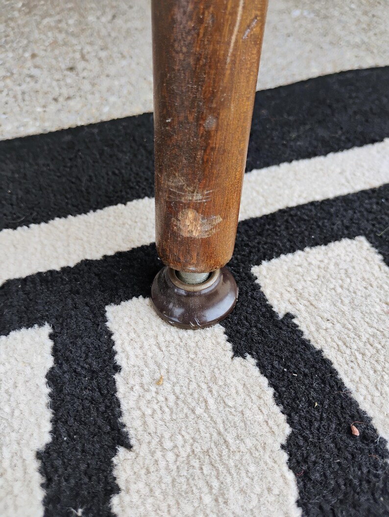 Puede incluir: Una pata de mueble cil&iacute;ndrica de madera con un rasgu&ntilde;o visible y un acabado desgastado cerca de la base, unida a un deslizador de suelo o tapa de pl&aacute;stico de color marr&oacute;n oscuro. La pata est&aacute; colocada verticalmente, mostrando la veta.