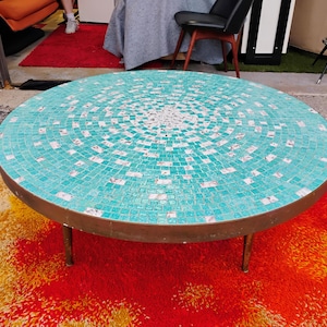 Op de afbeelding: Een ronde turquoise moza&iuml;ek salontafel met een messing onderstel en drie poten. De tafel staat op een rood en oranje shag tapijt.