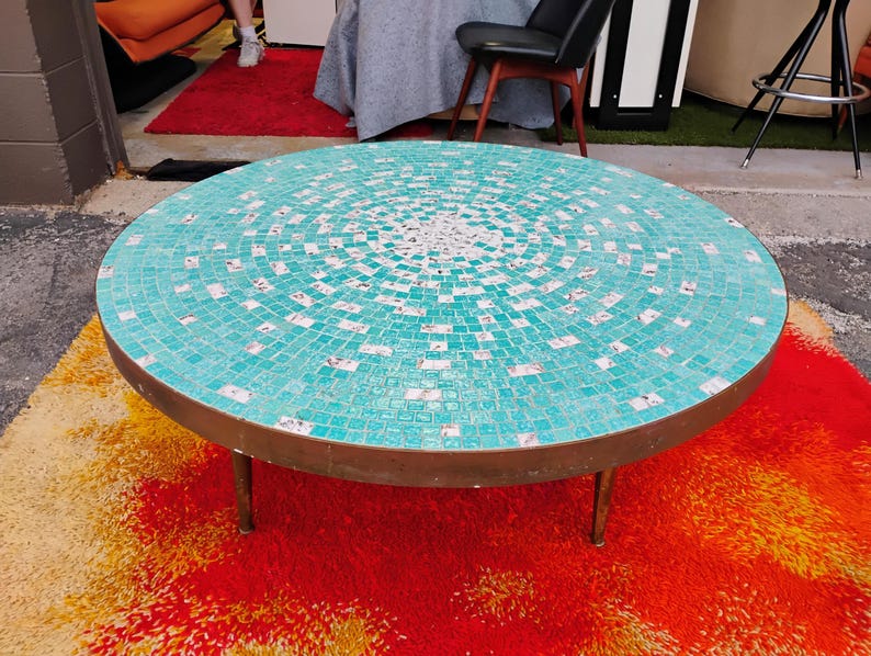 Op de afbeelding: Een ronde turquoise moza&iuml;ek salontafel met een messing onderstel en drie poten. De tafel staat op een rood en oranje shag tapijt.