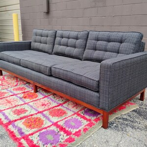 K&ouml;nnte beinhalten: Ein Dreisitzer-Sofa mit dunkelgrauem Stoffbezug und einem dezenten Karomuster. Das Sofa verf&uuml;gt &uuml;ber getuftete R&uuml;ckenkissen, gerade Armlehnen und einen Holzrahmen mit vier Beinen. Die Sitzkissen sind rechteckig und ebenfalls getuftet.