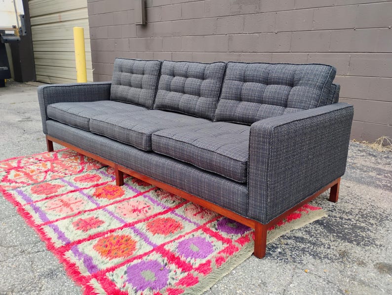 K&ouml;nnte beinhalten: Ein Dreisitzer-Sofa mit dunkelgrauem Stoffbezug und einem dezenten Karomuster. Das Sofa verf&uuml;gt &uuml;ber getuftete R&uuml;ckenkissen, gerade Armlehnen und einen Holzrahmen mit vier Beinen. Die Sitzkissen sind rechteckig und ebenfalls getuftet.