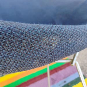 May include: A close-up view of a dark blue, textured fabric chair seat with a visible woven pattern. The chair features a metallic, silver-toned frame structure underneath the seat, showing a portion of the curved edge and the fabric's weave.