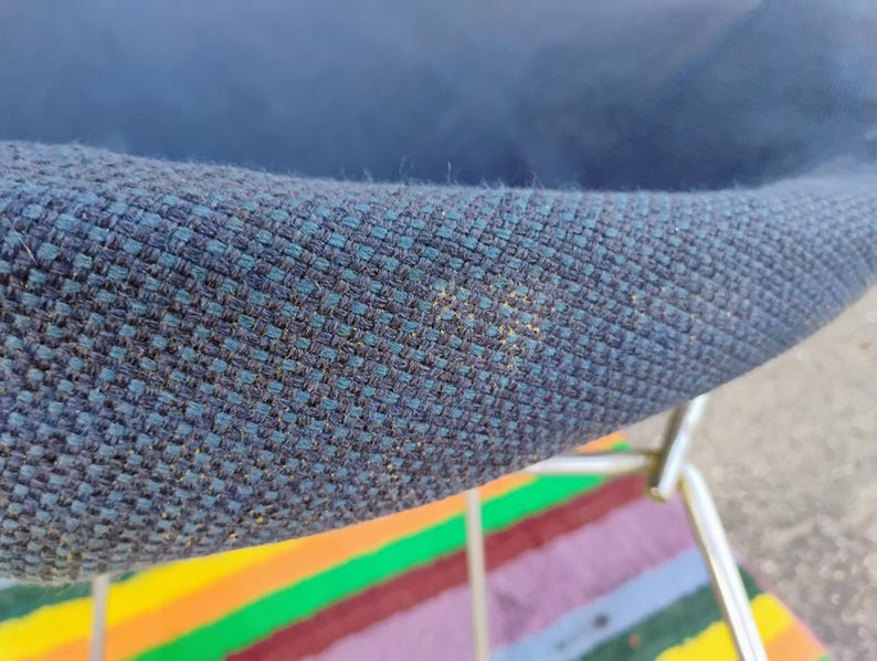 May include: A close-up view of a dark blue, textured fabric chair seat with a visible woven pattern. The chair features a metallic, silver-toned frame structure underneath the seat, showing a portion of the curved edge and the fabric's weave.