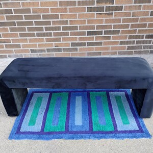 May include: A navy blue upholstered bench with a rectangular shape and curved edges. Beneath the bench is a rectangular rug with blue, green, and purple geometric patterns. The background is a brick wall.