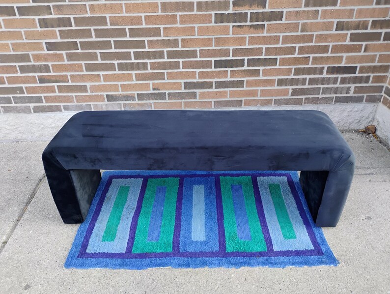 May include: A navy blue upholstered bench with a rectangular shape and curved edges. Beneath the bench is a rectangular rug with blue, green, and purple geometric patterns. The background is a brick wall.