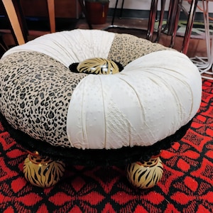 May include: A white and brown patterned round ottoman with a black trim and gold animal print legs. The ottoman has a tufted top with a large gold button in the center.