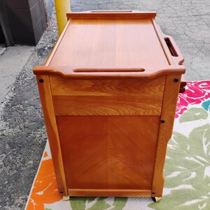 May include: A wooden serving cart with a top shelf and two drawers. The cart has a natural wood finish and is on wheels.