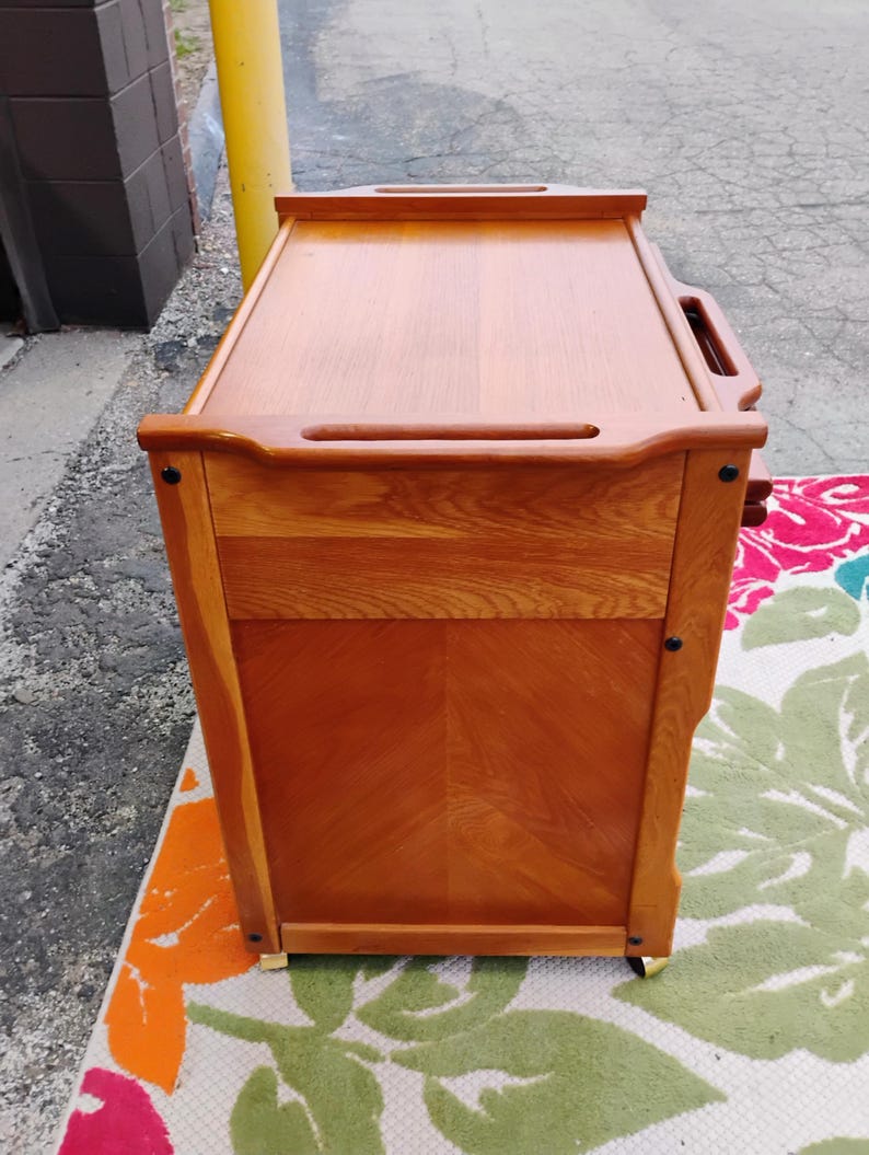 May include: A wooden serving cart with a top shelf and two drawers. The cart has a natural wood finish and is on wheels.