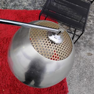 K&ouml;nnte beinhalten: Eine moderne, kugelf&ouml;rmige Lampe mit geb&uuml;rsteter Metalloberfl&auml;che. Die obere H&auml;lfte der Lampe hat einen perforierten Metallschirm. Ein Chromarm erstreckt sich von der Lampe. Die Lampe steht auf einem roten Teppich.