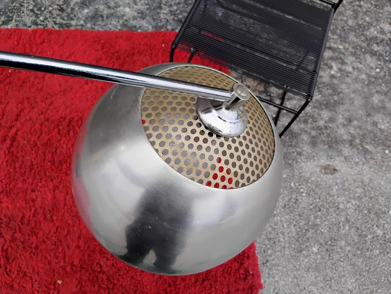 K&ouml;nnte beinhalten: Eine moderne, kugelf&ouml;rmige Lampe mit geb&uuml;rsteter Metalloberfl&auml;che. Die obere H&auml;lfte der Lampe hat einen perforierten Metallschirm. Ein Chromarm erstreckt sich von der Lampe. Die Lampe steht auf einem roten Teppich.