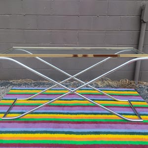 May include: A rectangular console table featuring a clear glass top with a polished gold-tone metal frame edge. The base consists of two intersecting chrome-finished metal legs that form an X-shape, providing a stable support structure.