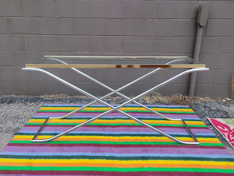 May include: A rectangular console table featuring a clear glass top with a polished gold-tone metal frame edge. The base consists of two intersecting chrome-finished metal legs that form an X-shape, providing a stable support structure.
