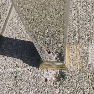K&ouml;nnte beinhalten: Eine klare Glasscheibe mit einem goldfarbenen, quadratischen Sockel. Die Scheibe ist abgewinkelt, und der Sockel ruht auf einer rauen, grauen Betonoberfl&auml;che. Das Bild ist bei nat&uuml;rlichem Licht aufgenommen und wirft einen Schatten.