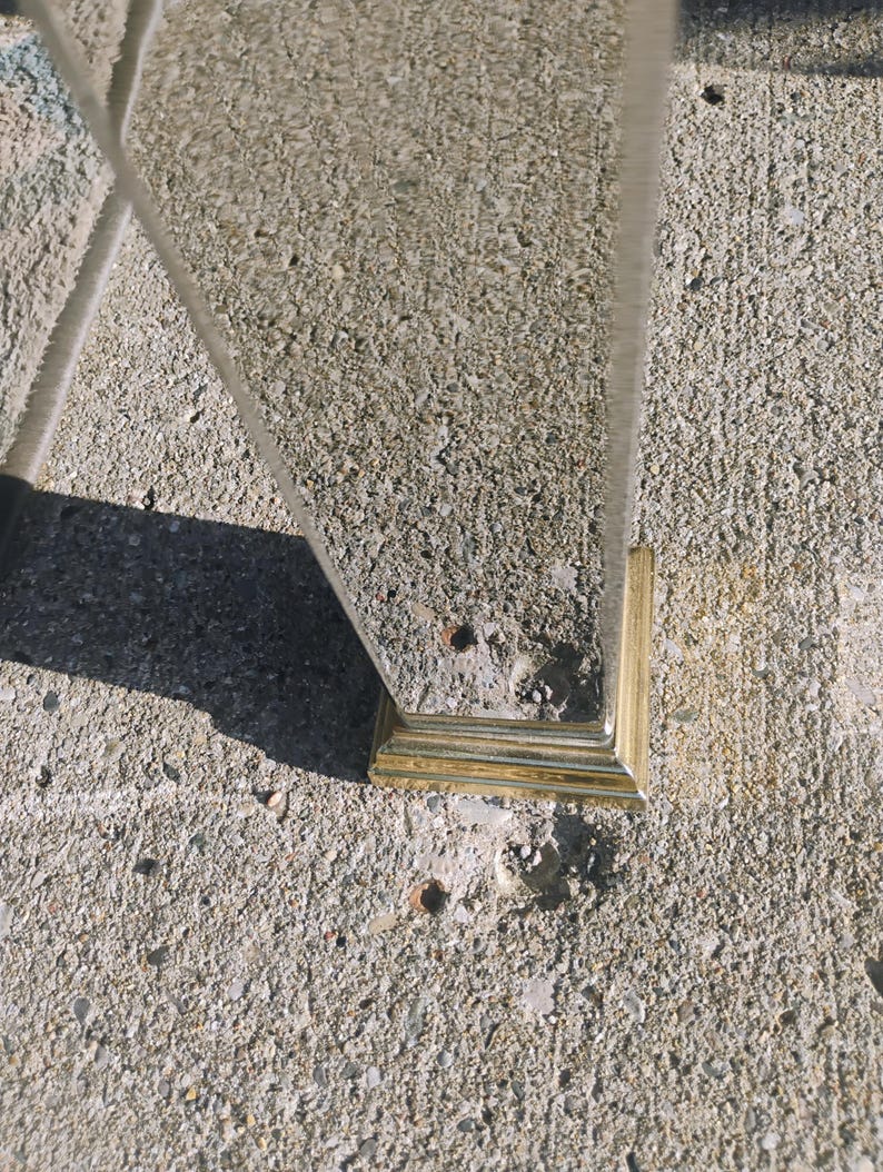 K&ouml;nnte beinhalten: Eine klare Glasscheibe mit einem goldfarbenen, quadratischen Sockel. Die Scheibe ist abgewinkelt, und der Sockel ruht auf einer rauen, grauen Betonoberfl&auml;che. Das Bild ist bei nat&uuml;rlichem Licht aufgenommen und wirft einen Schatten.