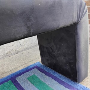 May include: A section of a dark gray upholstered furniture piece with a curved top and a rectangular base. A colorful rug with blue, green, and purple stripes is visible beneath the furniture.