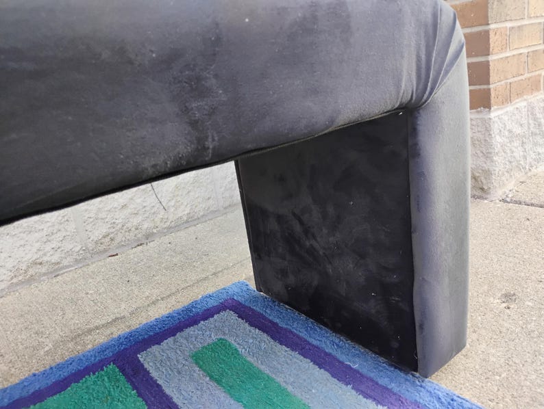 May include: A section of a dark gray upholstered furniture piece with a curved top and a rectangular base. A colorful rug with blue, green, and purple stripes is visible beneath the furniture.