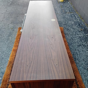 May include: A long, rectangular wooden sideboard with a dark brown wood grain finish. The top surface is flat and smooth, featuring a small circular light-colored inlay near the back edge and two small holes visible on the surface.