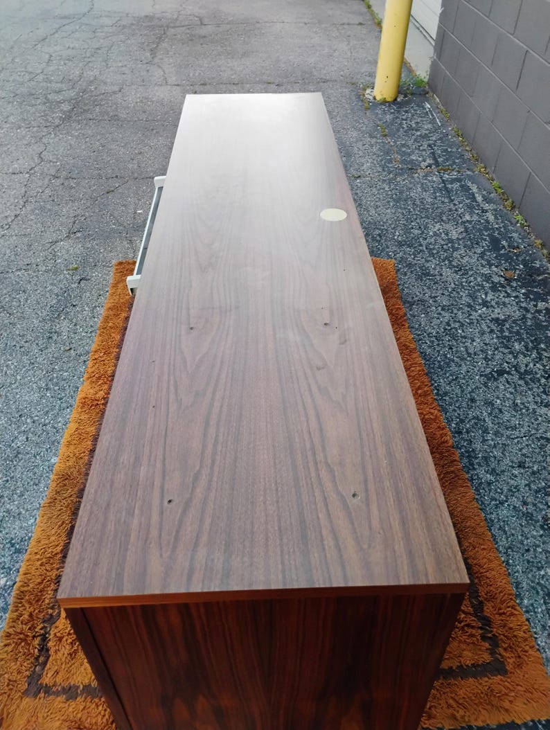 May include: A long, rectangular wooden sideboard with a dark brown wood grain finish. The top surface is flat and smooth, featuring a small circular light-colored inlay near the back edge and two small holes visible on the surface.