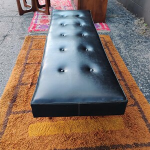 May include: A long, rectangular black bench featuring a tufted surface with ten evenly spaced buttons. The bench has a smooth, leather-like finish and a clean, straight-edged design, positioned horizontally to showcase its elongated, flat top surface.