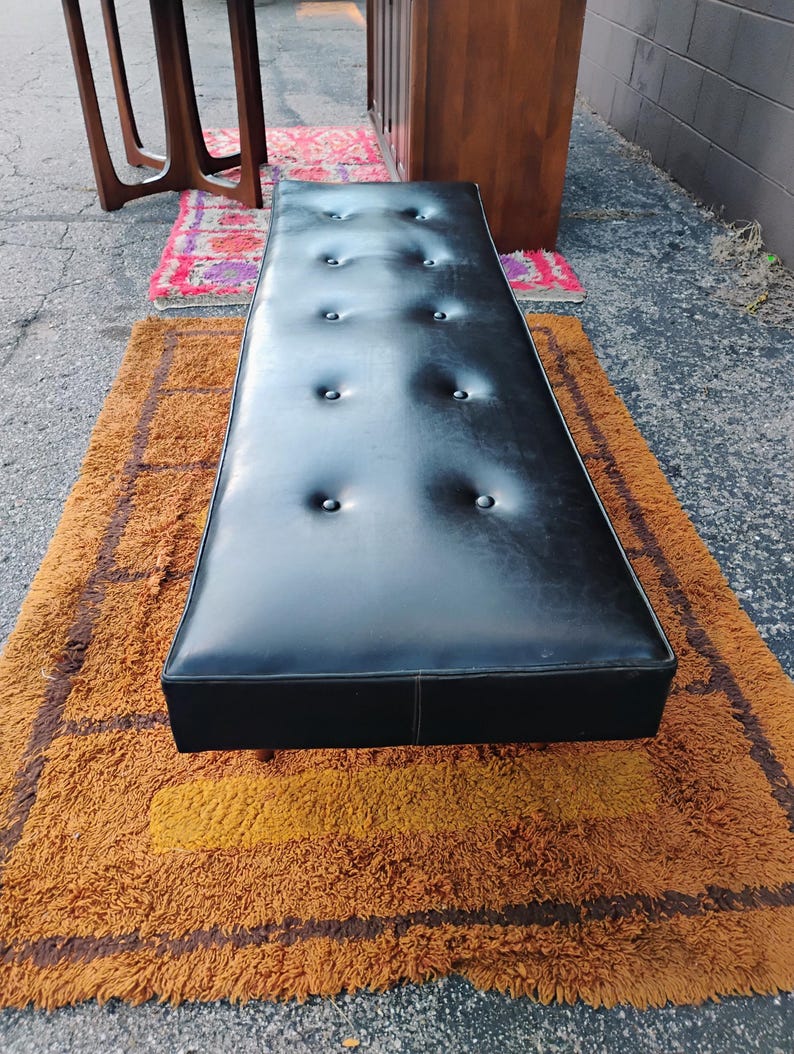 May include: A long, rectangular black bench featuring a tufted surface with ten evenly spaced buttons. The bench has a smooth, leather-like finish and a clean, straight-edged design, positioned horizontally to showcase its elongated, flat top surface.