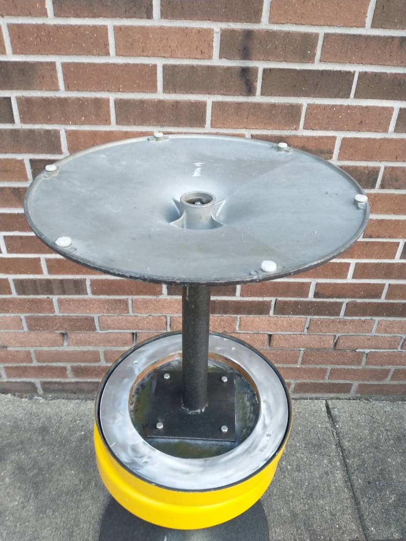 May include: A metal ashtray with a yellow base and a silver top. The top is round and has a funnel shape in the center. The base is a cylinder with a raised ring around the top.