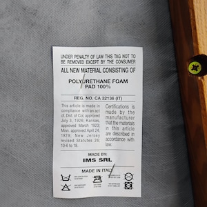May include: A white rectangular product tag attached to a gray fabric surface with a wooden frame edge. The tag features black text stating: UNDER PENALTY OF LAW THIS TAG NOT TO BE REMOVED EXCEPT BY THE CONSUMER, ALL NEW MATERIAL CONSISTING OF POLYURETHANE FOAM PAD 100%, REG. NO. CA 32136 (IT), MADE BY IMS SRL, MADE IN ITALY, and various care symbols.