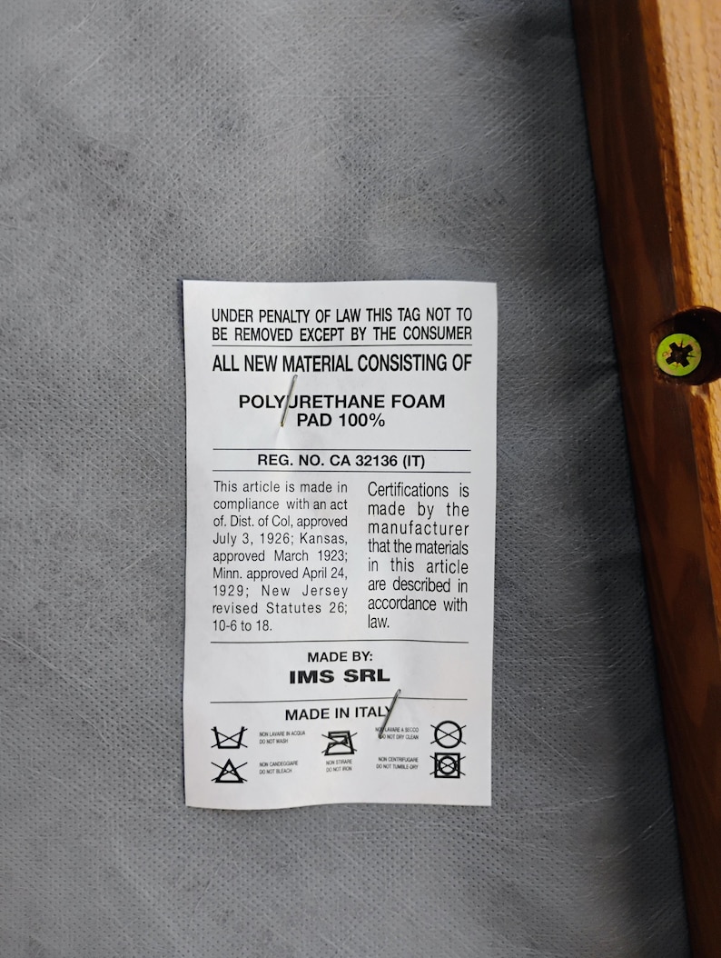 May include: A white rectangular product tag attached to a gray fabric surface with a wooden frame edge. The tag features black text stating: UNDER PENALTY OF LAW THIS TAG NOT TO BE REMOVED EXCEPT BY THE CONSUMER, ALL NEW MATERIAL CONSISTING OF POLYURETHANE FOAM PAD 100%, REG. NO. CA 32136 (IT), MADE BY IMS SRL, MADE IN ITALY, and various care symbols.