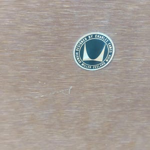 May include: A light brown wood surface featuring a circular black and silver metallic label. The text on the label reads: DESIGNED BY CHARLES EAMES HERMAN MILLER ZEELAND MICH. The label includes a stylized graphic of two curved shapes.