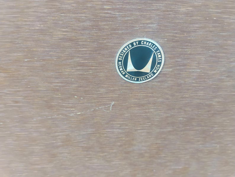 May include: A light brown wood surface featuring a circular black and silver metallic label. The text on the label reads: DESIGNED BY CHARLES EAMES HERMAN MILLER ZEELAND MICH. The label includes a stylized graphic of two curved shapes.