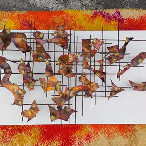 May include: Abstract wall art sculpture featuring a grid of metal pieces in shades of brown, orange, and red. The pieces are arranged in a random pattern and are attached to a white background.