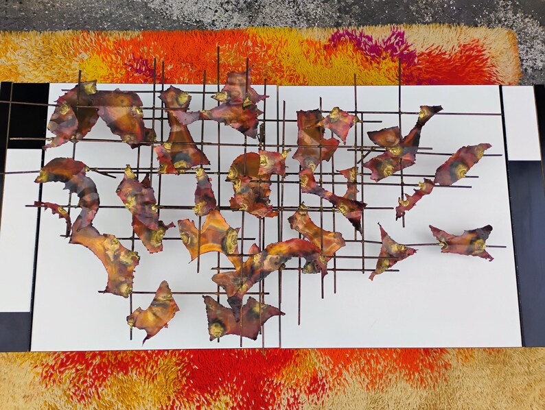 May include: Abstract wall art sculpture featuring a grid of metal pieces in shades of brown, orange, and red. The pieces are arranged in a random pattern and are attached to a white background.